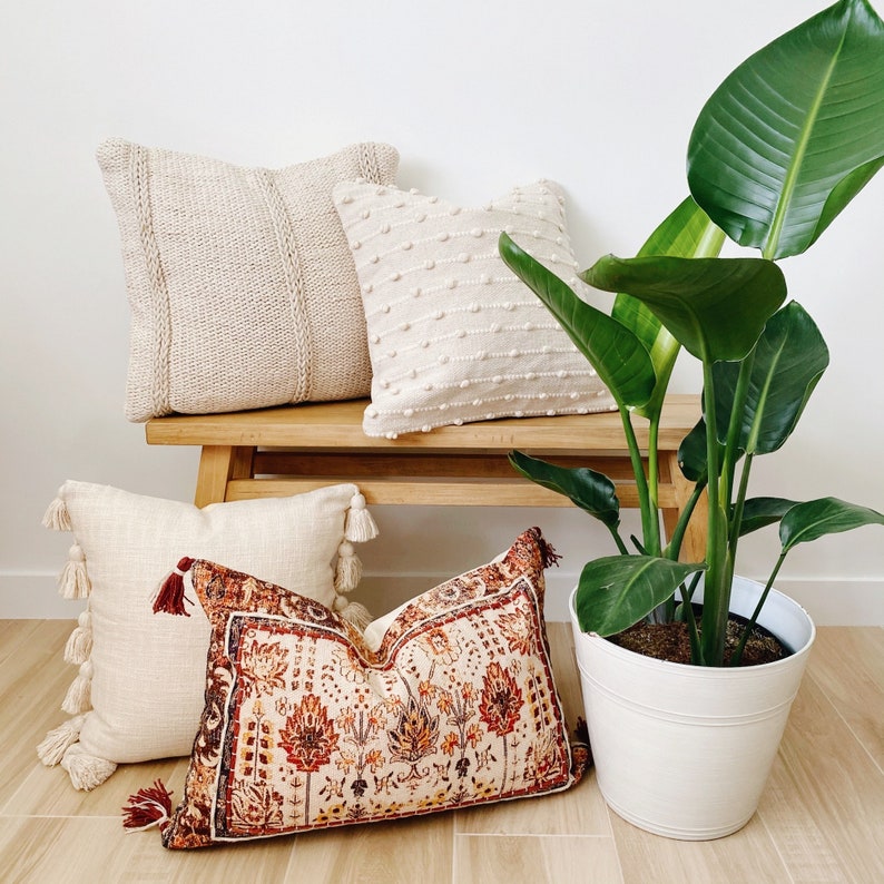 May include: A wooden bench with four throw pillows, two in a cream colour and two with a floral pattern. A large potted plant with green leaves is in front of the bench.