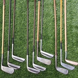 May include: A collection of vintage golf clubs with wooden shafts and leather grips, arranged on a green grassy surface. The clubs vary in color, with some showing signs of age and wear, and are positioned vertically.