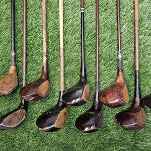 May include: A collection of antique golf clubs with wooden shafts and clubheads. The clubheads vary in color from light to dark brown, and the shafts are a mix of light and dark wood. The clubs are arranged on a green surface.