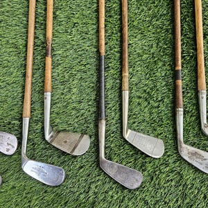 May include: A collection of eight vintage golf clubs featuring wooden shafts with dark grip sections and metallic heads. Each club displays unique wear, including scuffs and markings on the metal surfaces, arranged in a parallel vertical row.