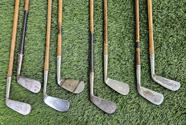 May include: A collection of eight vintage golf clubs featuring wooden shafts with dark grip sections and metallic heads. Each club displays unique wear, including scuffs and markings on the metal surfaces, arranged in a parallel vertical row.