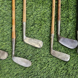 May include: A collection of eight vintage golf clubs featuring wooden shafts and metal heads. The clubs are arranged in a row, showcasing various head shapes and designs, including irons and putters with visible metal textures and aged wood.