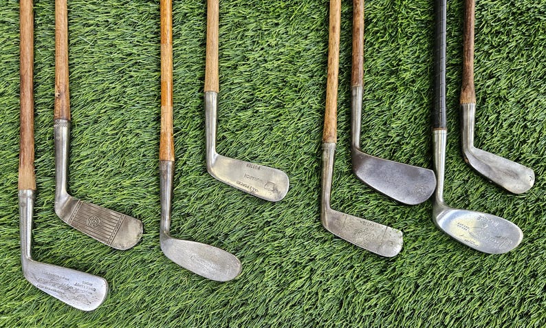 May include: A collection of eight vintage golf clubs featuring wooden shafts and metal heads. The clubs are arranged in a row, showcasing various head shapes and designs, including irons and putters with visible metal textures and aged wood.