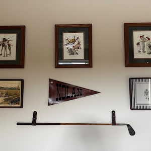 May include: A wall display featuring three framed vintage baseball prints, one framed baseball field illustration, one framed football player print, a triangular pennant with the text NOLAN, and a horizontal golf club mounted on two straps.
