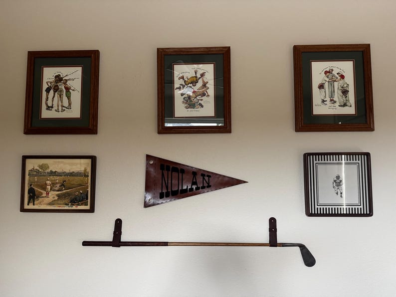 May include: A wall display featuring three framed vintage baseball prints, one framed baseball field illustration, one framed football player print, a triangular pennant with the text NOLAN, and a horizontal golf club mounted on two straps.