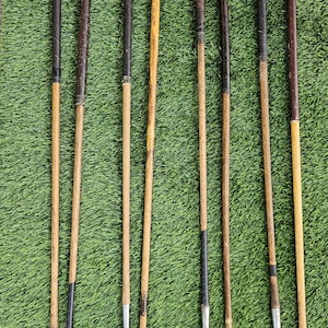 May include: A collection of seven vintage golf clubs featuring wooden shafts and metal clubheads. Each club has a dark leather or synthetic grip at the top, with varying wood grain patterns on the shafts and distinct metal head designs.