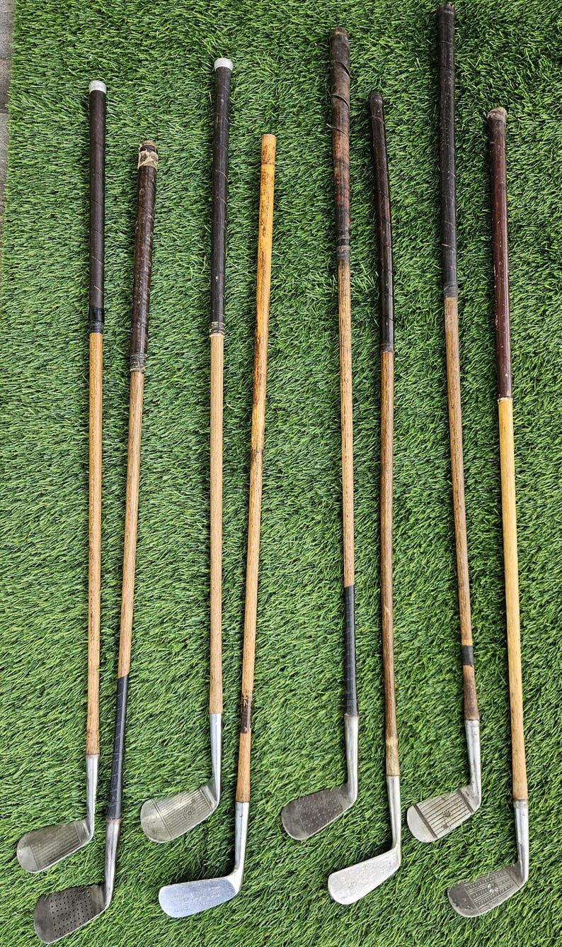 May include: A collection of seven vintage golf clubs featuring wooden shafts and metal clubheads. Each club has a dark leather or synthetic grip at the top, with varying wood grain patterns on the shafts and distinct metal head designs.