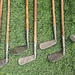 May include: A collection of eight vintage golf clubs featuring wooden shafts and metal heads. The clubs display various head shapes, including irons and putters, with textured faces and metallic finishes. The wooden shafts show natural grain patterns.