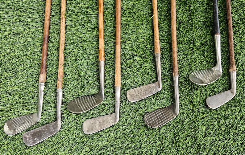 May include: A collection of eight vintage golf clubs featuring wooden shafts and metal heads. The clubs display various head shapes, including irons and putters, with textured faces and metallic finishes. The wooden shafts show natural grain patterns.