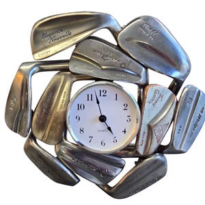 May include: A circular wall clock featuring a white face with black numerals and hands, surrounded by a decorative frame constructed from various silver-toned metal golf club heads arranged in a radial pattern, with the word Quartz printed on the face.