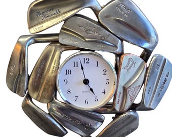 May include: A circular wall clock featuring a white face with black numerals and hands, surrounded by a decorative frame constructed from various silver-toned metal golf club heads arranged in a radial pattern, with the word Quartz printed on the face.