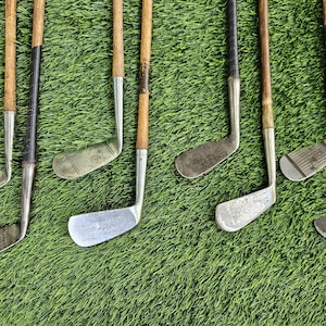 May include: A collection of eight vintage golf clubs featuring wooden shafts and metal clubheads. The clubs display various head shapes, including irons and putters, with some showing textured faces and dark leather or tape grips on the handles.