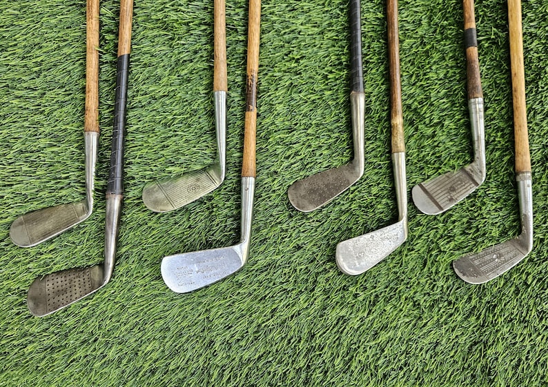 May include: A collection of eight vintage golf clubs featuring wooden shafts and metal clubheads. The clubs display various head shapes, including irons and putters, with some showing textured faces and dark leather or tape grips on the handles.