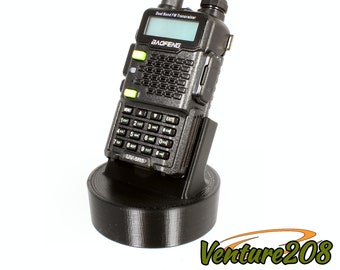 May include: A black handheld two-way radio with a green display screen and a black charging base. The radio has a keypad with numbers and a few buttons. The radio is labeled "BAOFENG" and "UV-5R5".
