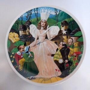 May include: A decorative plate featuring a scene from The Wizard of Oz. The plate depicts the Good Witch in a pink gown, surrounded by characters. The background includes a landscape with buildings and trees. The plate has a white border.
