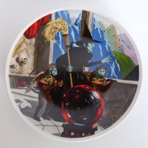 May include: A decorative plate featuring a scene from The Wizard of Oz. The image depicts the Wicked Witch of the West, two winged monkeys, and a crystal ball. The background includes a window with a mountain view and a red curtain.