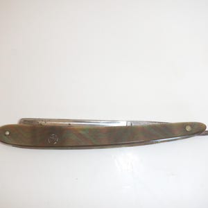 May include: A straight razor with a folded metal blade and a handle featuring a mottled, wavy pattern in shades of green, brown, and gold. The handle is secured with two metal pins and displays a small circular logo on the side surface.