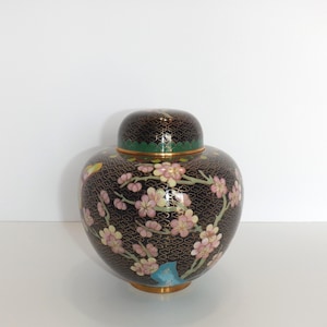 May include: A black cloisonné jar with a floral design. The jar is decorated with pink and white flowers, green leaves, and gold accents. The lid is also decorated with a floral design.