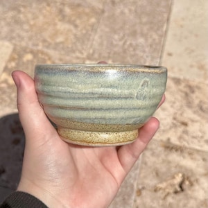 May include: A handmade ceramic bowl with a light green and brown glaze. The bowl has a rounded shape with horizontal ridges and a small, circular base. The bowl is held in a hand, showcasing its size and texture.
