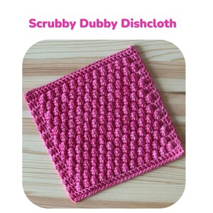 May include: A bright pink, square dishcloth with a textured, bumpy surface. The words "Scrubby Dubby Dishcloth" are above the cloth. The dishcloth is made with cluster stitches, creating a scrubby texture. The text below says, "This dishcloth uses cluster stitches to form a texture that creates something scrubby dubby!"
