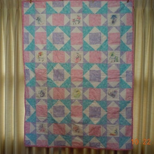 Könnte beinhalten: Eine rosafarbene und blaue Patchwork-Decke mit weißem Hintergrund. Die Decke besteht aus Quadraten mit verschiedenen Designs, darunter Sterne, Blumen und Tiere. Die Decke hängt an einer Wand.