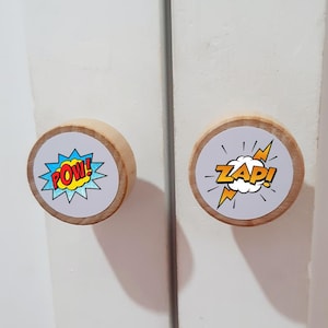 Pode incluir: Dois botões de armário de madeira com etiquetas redondas brancas. O botão da esquerda tem um gráfico POW! de estilo de banda desenhada azul e amarelo. O botão da direita tem um gráfico ZAP! de estilo de banda desenhada azul e amarelo.