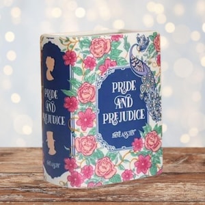 Può includere: Un contenitore decorativo a forma di libro con il titolo "Pride and Prejudice" sul davanti. Il design presenta un motivo floreale con rose rosa e un pavone. Lo sfondo è un effetto bokeh sfocato.