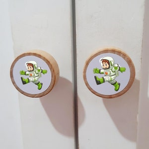 May include: Two round wooden cabinet knobs featuring a light-colored natural wood edge and a central circular graphic. The graphic depicts a cartoon astronaut in a white and green space suit, shown in a running pose against a light gray background.