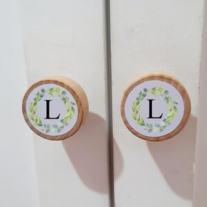 Puede incluir: Dos pomos de armario redondos de madera con una incrustación circular blanca. Cada pomo muestra una letra L negra centrada dentro de un diseño de corona de hojas verdes impreso al estilo acuarela. Montados en una puerta blanca.