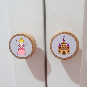 Può includere: Due pomelli rotondi in legno per armadi fissati su ante bianche. Il pomello sinistro presenta una principessa dei cartoni animati in un abito rosa con cuori, mentre quello destro mostra un castello beige con torri viola su sfondo bianco.