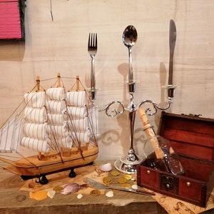 Puede incluir: Un velero de madera con velas blancas se encuentra sobre una superficie de madera con un candelabro de plata, un tenedor, una cuchara, una botella con un mensaje dentro y un cofre del tesoro de madera.