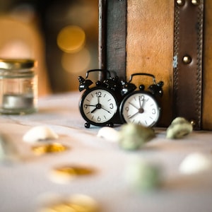 Könnte beinhalten: Zwei kleine schwarze Wecker mit weißen Zifferblättern und schwarzen Zeigern stehen auf einem weißen Tisch. Die Wecker sind scharf gestellt, während der Hintergrund unscharf ist. Um die Wecker herum liegen kleine Dekorationsgegenstände.