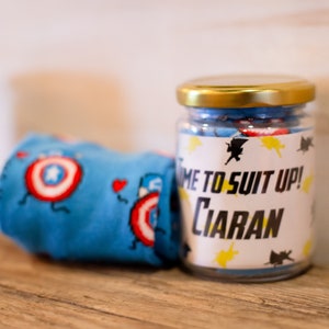 May include: A glass jar with a gold lid filled with blue and red candies. The jar has a label that reads "TIME TO SUIT UP! Ciaran" with superhero graphics. A blue fabric item with Captain America shield designs is next to the jar.
