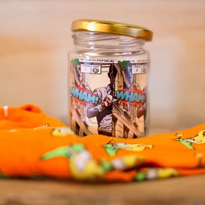 May include: A clear glass jar with a gold lid and a comic book design featuring a superhero with the words "WHAM!" printed on the jar. The jar is sitting on top of a pair of orange socks with a yellow and green pattern.