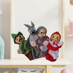 Peut inclure: Un ensemble de marionnettes faites à la main, dont un personnage vêtu de vert, un âne gris, une vieille femme et une fille en cape rouge. Les marionnettes sont disposées devant un cadre photo vierge. Une grande girafe blanche est à droite.