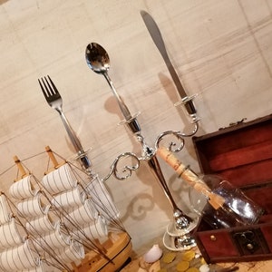 Puede incluir: Un modelo de barco de madera con velas blancas, un tenedor, una cuchara y un cuchillo de plata, un candelabro de plata, una botella de vidrio con un papel enrollado dentro y un cofre del tesoro de madera con un interior marr&oacute;n.