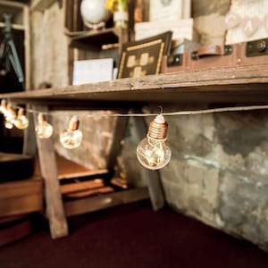 Op de afbeelding: Een rustieke houten tafel met een string van warm witte lampjes die er vanaf hangen. De lampjes zijn in de vorm van kleine, heldere glazen bollen met koperen accenten. De tafel is versierd met vintage koffers en andere items.