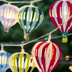 Könnte beinhalten: Dekorative Lichterkette mit bunten Heißluftballons, gestreift in Rot und Weiß, Grün und Weiß sowie Gelb und Weiß. Die Lichter sind an einem dünnen weißen Draht aufgereiht und strahlen ein warmes Licht aus. Ideal für Dekorationen.