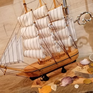 Puede incluir: Un modelo de velero de madera con velas blancas y un casco de madera marr&oacute;n. El velero est&aacute; sentado sobre una superficie de madera con conchas marinas, monedas de oro y un tenedor.