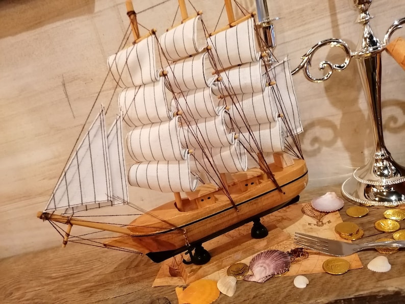 Puede incluir: Un modelo de velero de madera con velas blancas y un casco de madera marr&oacute;n. El velero est&aacute; sentado sobre una superficie de madera con conchas marinas, monedas de oro y un tenedor.