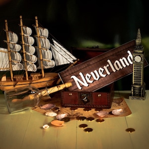 Puede incluir: Un cofre del tesoro de madera con un letrero que dice "Neverland" en la parte superior. También hay un pequeño barco de modelo, una botella de vidrio con un corcho y un Big Ben en miniatura en la mesa. Hay monedas de oro y conchas esparcidas alrededor del cofre.