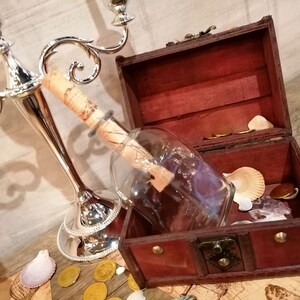 Puede incluir: Un cofre del tesoro de madera con un cierre de lat&oacute;n est&aacute; abierto, revelando una botella de vidrio con un trozo de papel enrollado en su interior. La botella est&aacute; encima de algunas monedas de oro y conchas marinas.