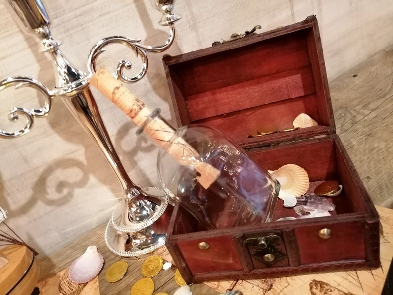 Puede incluir: Un cofre del tesoro de madera con un cierre de lat&oacute;n est&aacute; abierto, revelando una botella de vidrio con un trozo de papel enrollado en su interior. La botella est&aacute; encima de algunas monedas de oro y conchas marinas.