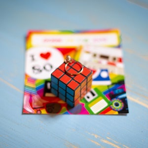 Può includere: Un portachiavi a forma di cubo di Rubik colorato con un anello dorato. Il portachiavi è appoggiato su una carta con il testo "I Love 80s" e altre grafiche a tema anni '80.