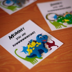 Könnte beinhalten: Eine weiße Karte mit dem Text "Mummy you are rawwwwsome!" und einem blauen Dinosaurier-Schlüsselanhänger mit gelben Akzenten. Die Karte liegt auf einer Holzoberfläche.