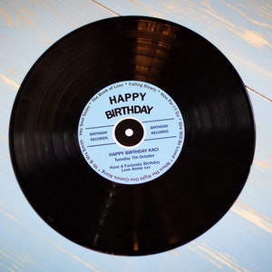 Op de afbeelding: Een zwarte vinylplaat met een lichtblauw label met de tekst "HAPPY BIRTHDAY". Het label bevat ook de tekst "BIRTHDAY RECORDS" en een gepersonaliseerde boodschap. De plaat ligt op een lichtblauwe houten achtergrond.