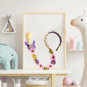 Könnte beinhalten: Ein dekoratives Stirnband mit einem lila Band und einer kleinen Krone, verziert mit rosa und gelben Blumen. Ein blondes Zopf mit einer lila Schleife ist befestigt. Das Stirnband wird in einem Holzrahmen ausgestellt.