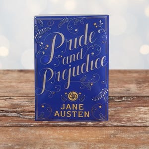 Può includere: Un libro con copertina rigida "Pride and Prejudice" di Jane Austen. La copertina è blu scuro con scritte dorate e decorazioni floreali. Il libro è appoggiato su una superficie di legno.