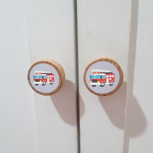May include: Two round wooden cabinet knobs featuring a graphic illustration of a red fire truck with a white ladder on top. The knobs have a light natural wood finish on the sides and a flat circular face with a light gray background.