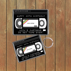 May include: A black and silver cassette tape keychain with the text "Insert this side into recorder" and "Happy 30th Birthday" on the front. The cassette tape has the text "AZZA'S CARTOONS" and "VHS" on it. The text "DO NOT TAPE OVER!" is printed below the cassette tape.